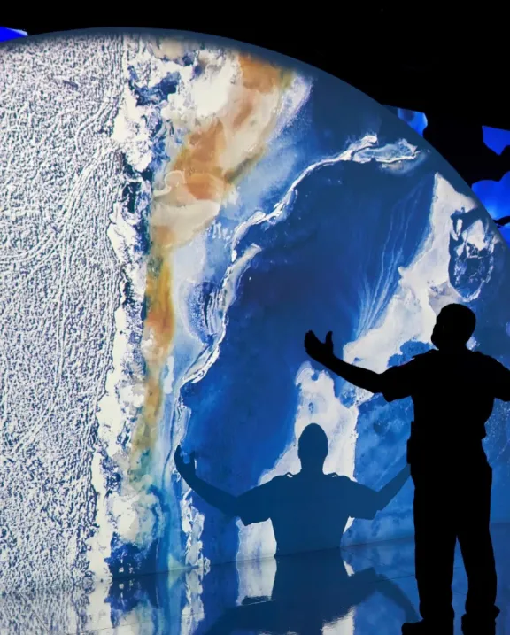 Projection of a planet with a silhouette of a man with his hands in the air looking at a large projection of a planet